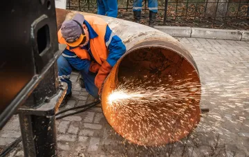 Рабочий режет металлическую трубу теплотрассы болгаркой на фоне спора о сносе гаражей в Киеве ради деревьев