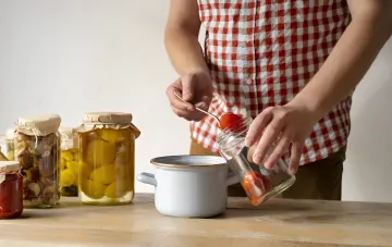 Человек в красно-белой клетчатой рубашке аккуратно выкладывает помидоры из стеклянной банки в эмалированную кастрюлю, на столе стоят заготовки в банках с грибами и желтыми овощами, сезон заготовок 2026