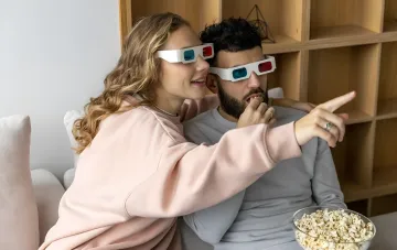Молодая пара в очках 3D смотрит фильм на диване, наслаждаясь попкорном, иллюстрируя тему ленивости и любви к домашнему отдыху