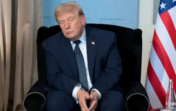 Дональд Трамп сидит в кресле с серьезным выражением лица, рядом американский флаг, что символизирует его текущие политические трудности.