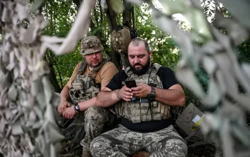 Украинские военные в камуфляже в лесу, один из них смотрит в смартфон, обсуждая стратегию в ответ на угрозы РФ