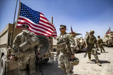 Американские солдаты в камуфляже с оружием и флагом США на фоне военной техники — иллюстрация к новости о том, что Иран объявил «охоту» на них с наградой $30 000