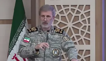 Iranian military officer in camouflage uniform delivers a speech in front of the Iranian flag, showing determination in response to US threats regarding the Strait of Hormuz.