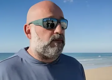 Man with beard and sunglasses on the background of Cyprus beach, reflection in glasses shows people discussing the topic of antisemitism.