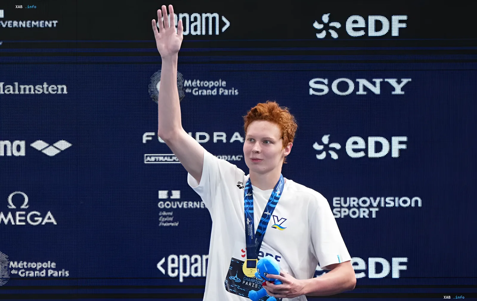 Никита Шеремет на пьедестале с золотой медалью чемпионата Европы по плаванию, машет рукой и держит сувенир