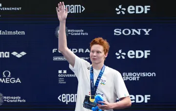 Никита Шеремет на пьедестале с золотой медалью чемпионата Европы по плаванию, машет рукой и держит сувенир