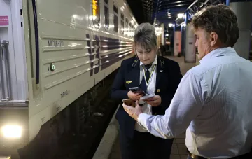 A woman in a railway uniform checks a passenger's documents on the platform next to a train, illustrating ticket verification and name transliteration for international travel.