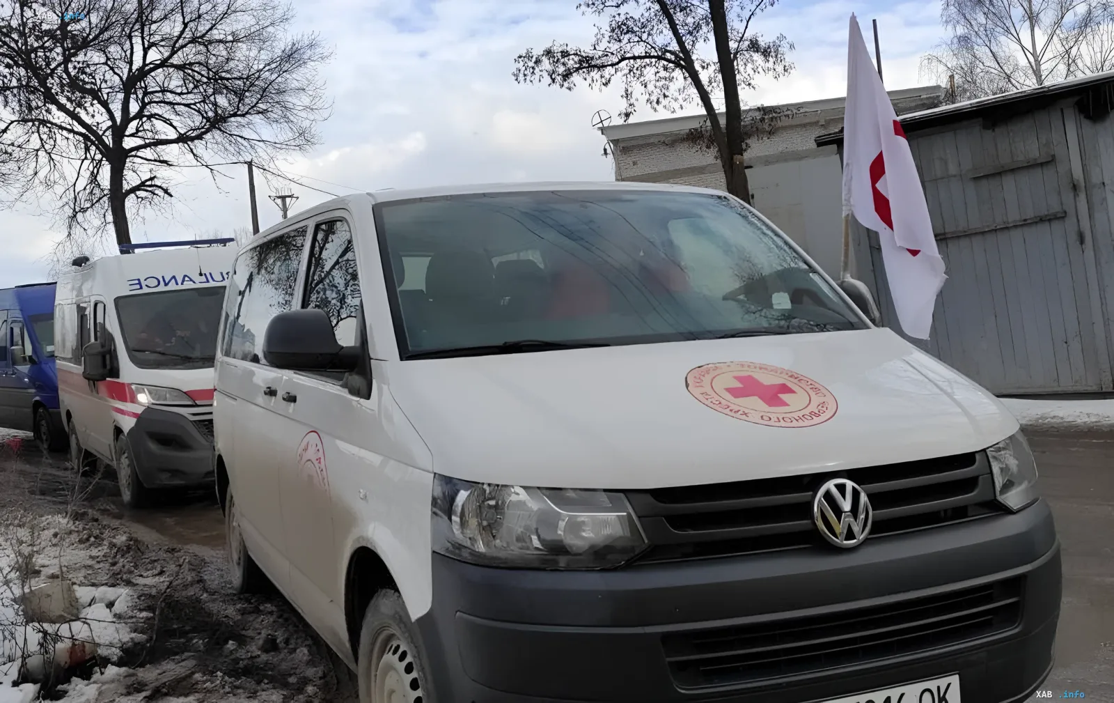 Белый фургон Volkswagen с эмблемой Украинского Красного Креста и красным крестом на капоте, припаркованный рядом с другим гуманитарным транспортом на фоне зимнего пейзажа.