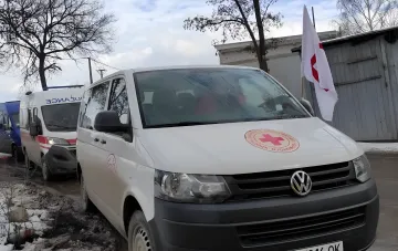 Белый фургон Volkswagen с эмблемой Украинского Красного Креста и красным крестом на капоте, припаркованный рядом с другим гуманитарным транспортом на фоне зимнего пейзажа.