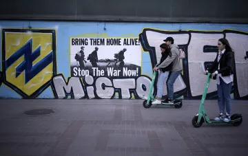Junge Menschen auf E-Scootern fahren an einem Graffiti mit der Aufschrift „Stop The War Now!“ in Kiew vorbei – ein Symbol der aktuellen Debatte über ein Verbot von E-Scootern nach tragischen Unfällen