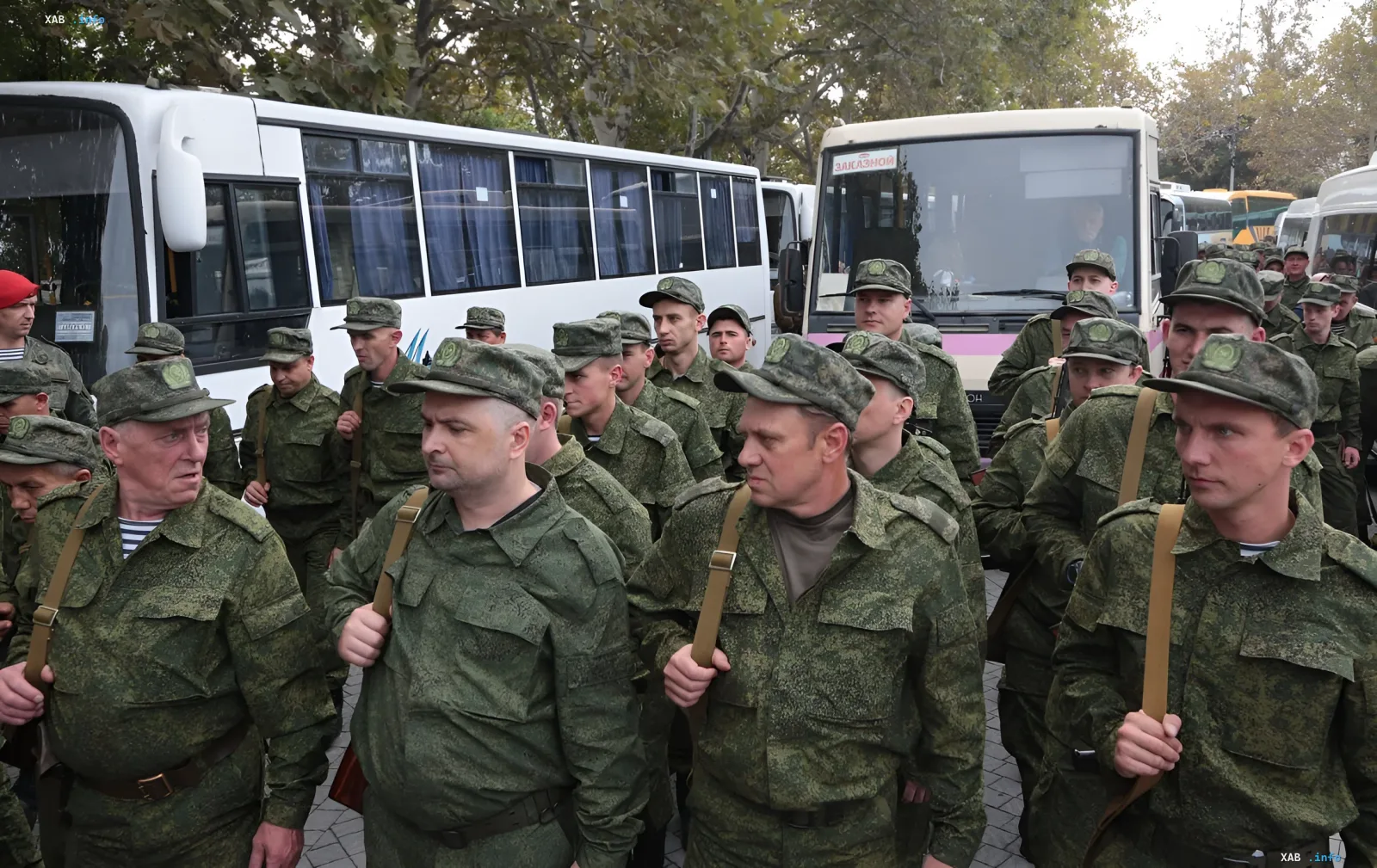 Группа российских военных в камуфляжной форме и фуражках с эмблемами, идущих рядом с белыми автобусами, иллюстрируя тему скрытых ограничений на выезд мужчин призывного возраста в России.