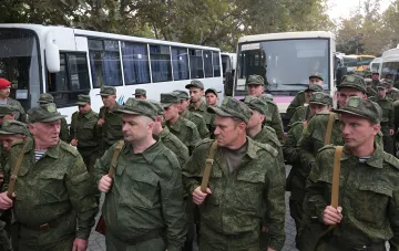 Группа российских военных в камуфляжной форме и фуражках с эмблемами, идущих рядом с белыми автобусами, иллюстрируя тему скрытых ограничений на выезд мужчин призывного возраста в России.