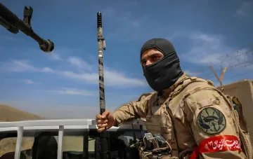 Военнослужащий в камуфляже и балаклаве с оружием на фоне военной техники, символизирующий угрозу со стороны Ирана