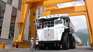 Огромный белый электрический самосвал Zijin Longking под жёлтым краном на заводе в Китае, рядом рабочие в касках