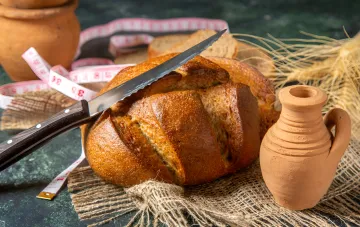Свежеиспеченная буханка хлеба без замеса с хрустящей корочкой, нарезанная ножом, рядом с глиняной посудой и пшеницей.