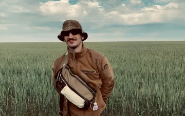 Віталій Капранов, барабанщик гурту «Квіткіс», на фоні зеленого поля у військовій формі з українським прапором на рукаві