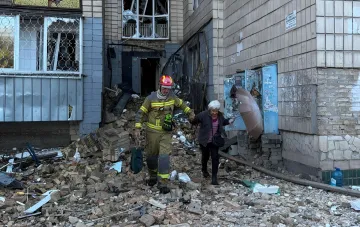 Пожарный помогает пожилой женщине пройти через завалы разрушенного жилого дома после комбинированной атаки на Киев ночью 20 августа