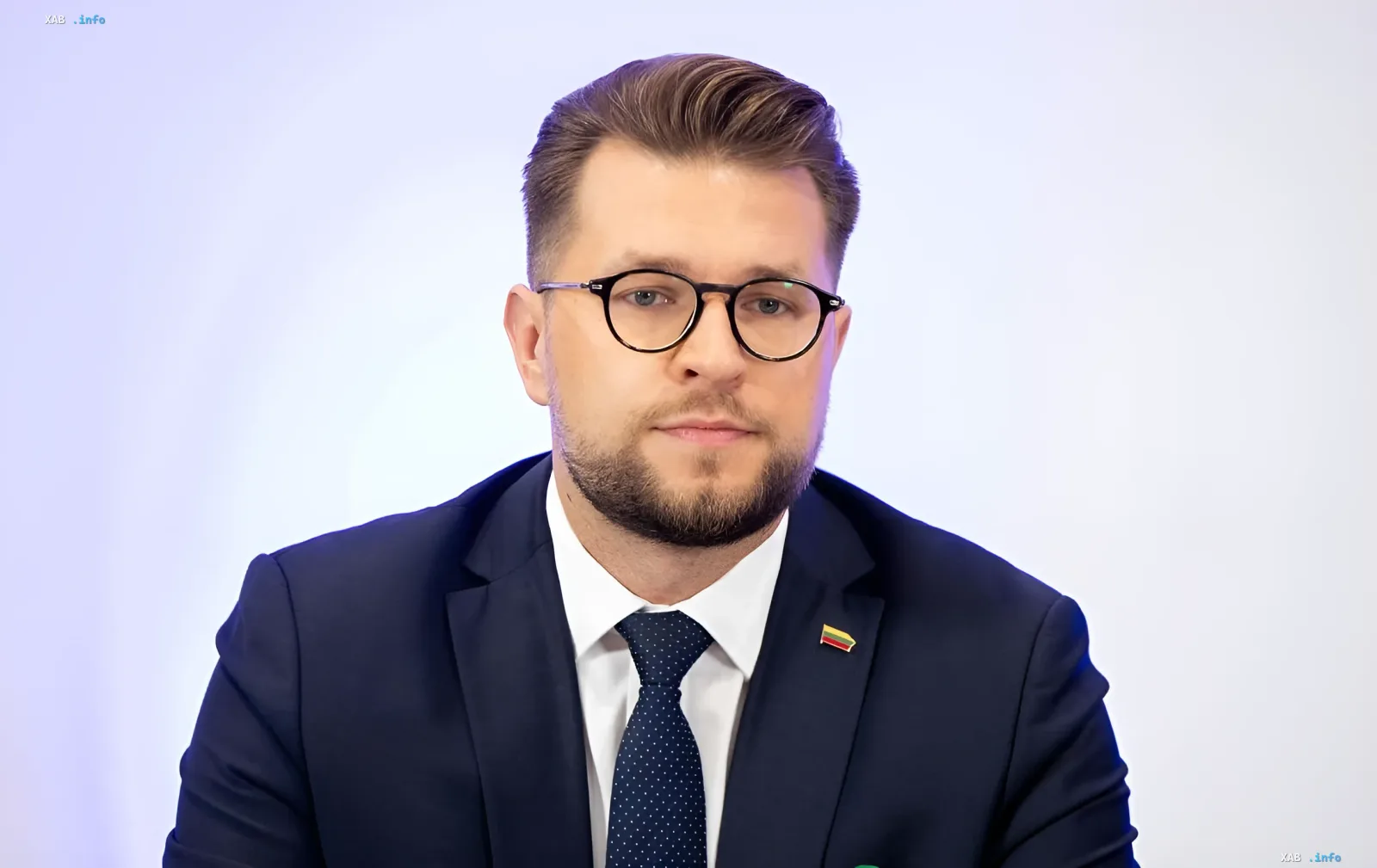 Министр энергетики Литвы в костюме с значком литовского флага на лацкане на фоне светлой стены