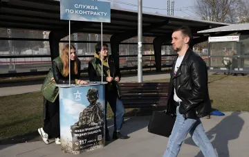 Агитационный пункт «Служба по контракту» Армии России на железнодорожной станции, где девушки в камуфляже раздают листовки, а мужчина с чемоданом проходит мимо — визуальный элемент негласного фильтра на границе