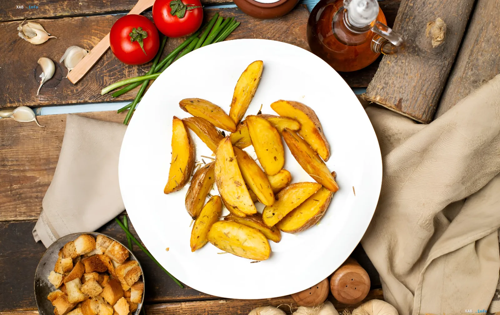 Запечённый картофель по-селянски дольками с хрустящей корочкой на белой тарелке рядом с помидорами, чесноком и зелёным луком