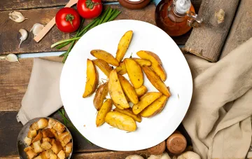 Запечённый картофель по-селянски дольками с хрустящей корочкой на белой тарелке рядом с помидорами, чесноком и зелёным луком
