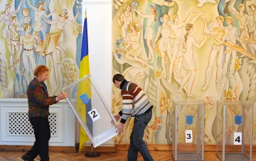 Голосование на выборах в Украине: люди переносят урну для голосования с гербом на фоне флага и исторической мозаики — выборы во время войны