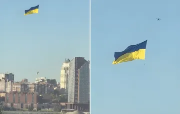 Гигантский сине-жёлтый флаг Украины парит в небе над Днепром в Киеве, рядом летит дрон-квадрокоптер, на заднем плане — городская застройка и набережная