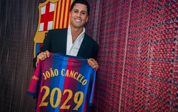 Жоау Канселу в костюме держит форму «Барселоны» с надписью 2029 на фоне эмблемы клуба после подписания контракта