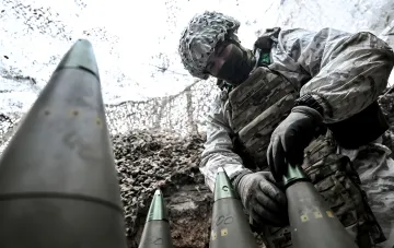Военнослужащий Сил обороны Украины в камуфляже готовит к применению дальнобойные 155-мм артиллерийские снаряды, иллюстрируя проблему дефицита боеприпасов