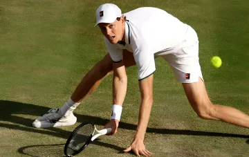 Jannik Sinner in white outfit on a grass court in a low, intense stance, preparing to hit the ball