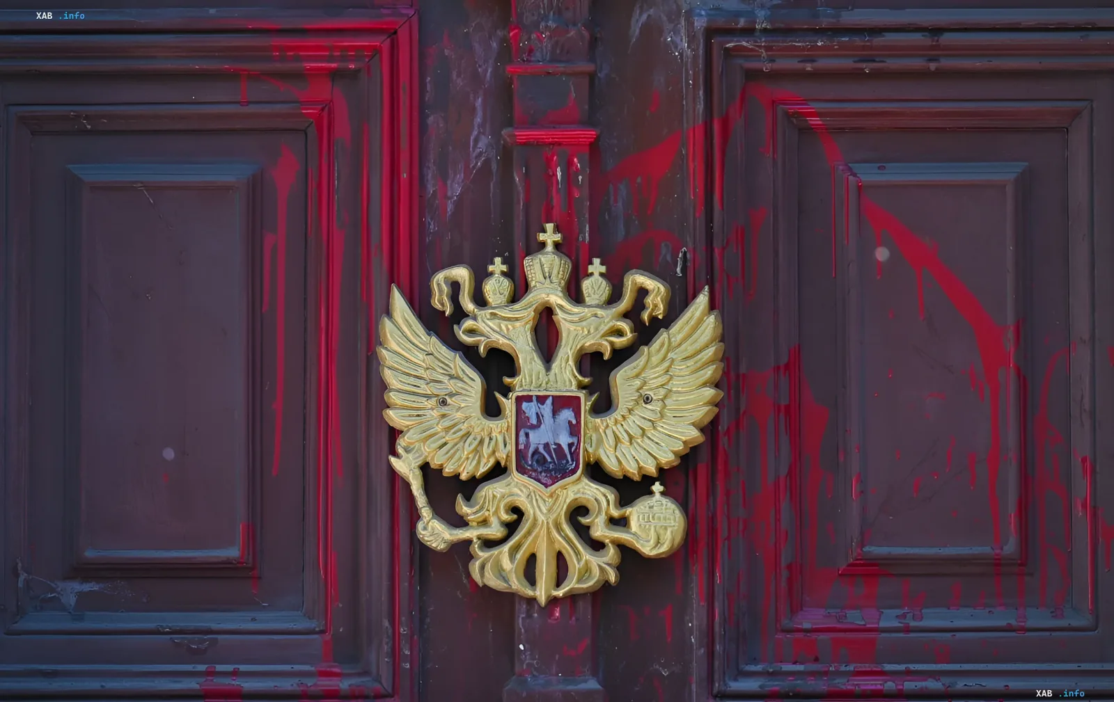 Золотой герб России — двуглавый орёл — на двери здания посольства на фоне красных брызг краски