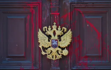 Золотой герб России — двуглавый орёл — на двери здания посольства на фоне красных брызг краски