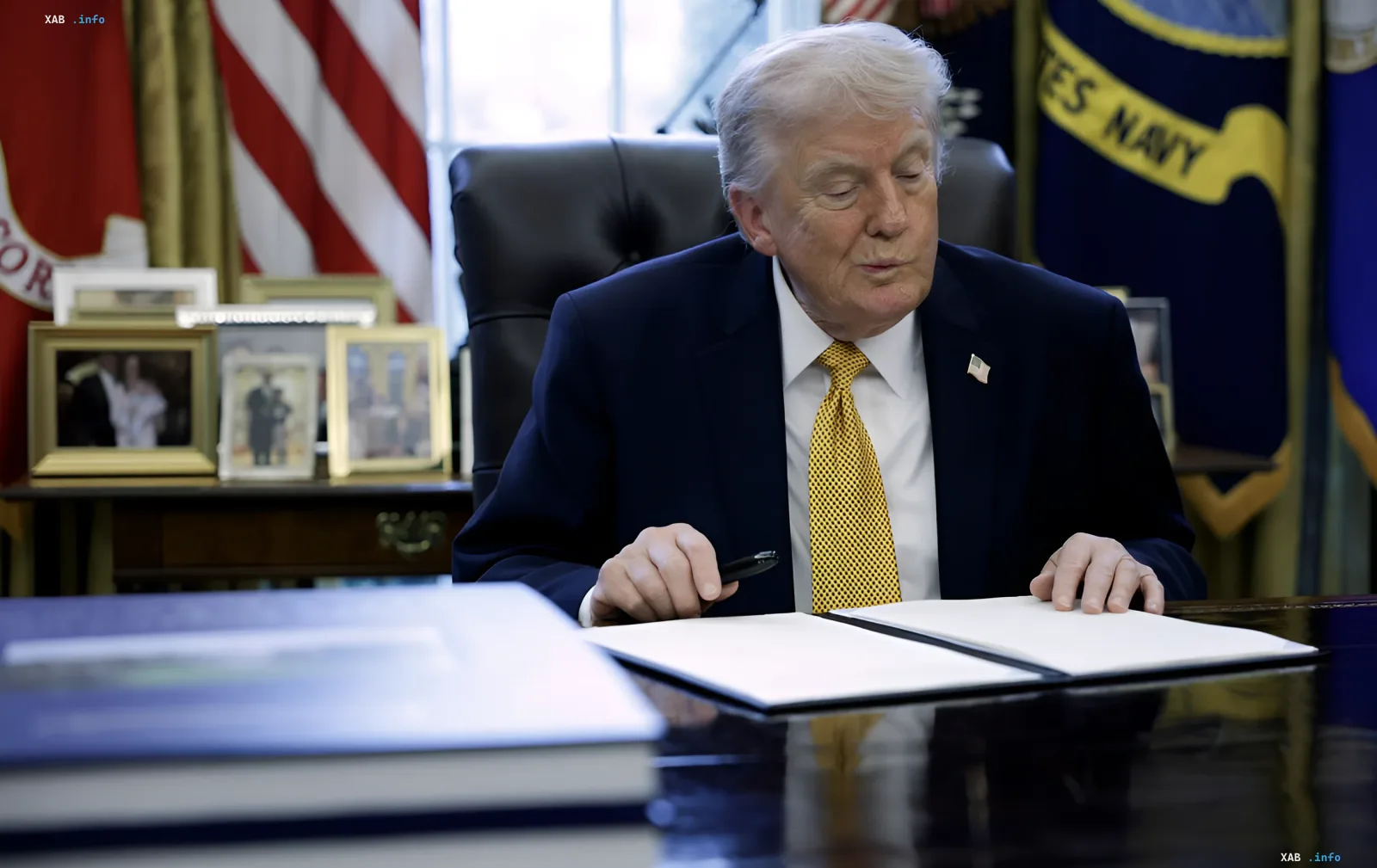 Donald Trump seated at the Resolute Desk in the Oval Office holding a pen over documents, US flags in the background