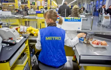 Кассир в синем жилете Metro за кассой Cash & Carry сканирует товары на фоне очереди покупателей в супермаркете