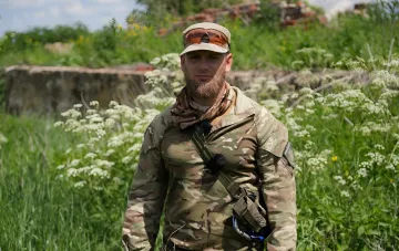 Suchteammitglied Alexey Yukov in Tarnkleidung vor dem Hintergrund zerstörter Gebäude und Wildblumen