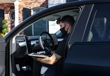 Водитель в маске и перчатках работает на ноутбуке в салоне электромобиля Tesla, в то время как салонная камера фиксирует его действия