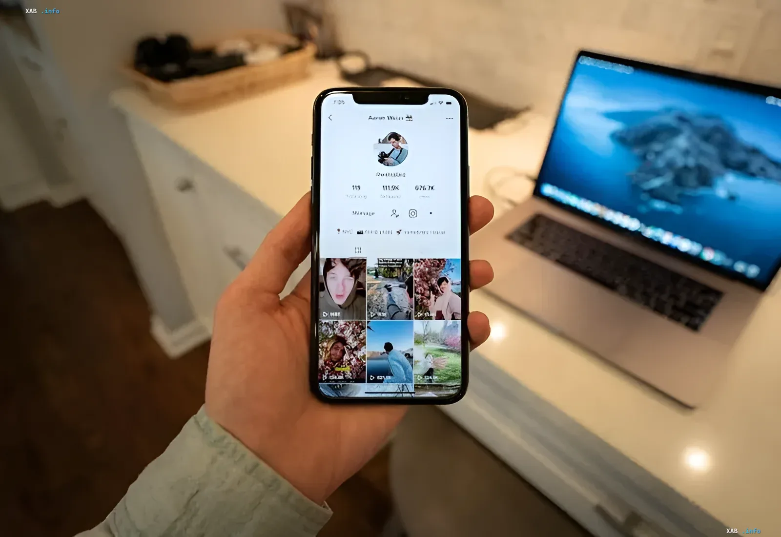A hand holding a smartphone displaying a TikTok profile with a laptop in the background