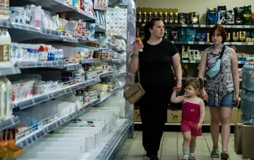 Покупательница с ребёнком выбирает продукты в проходе украинского супермаркета рядом с холодильной витриной и полками с базовыми товарами