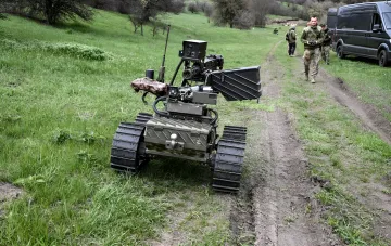 Geländegestützter roboterisierter Komplex auf Kettenlaufwerk mit Kamera und Manipulator, Bediener mit Fernsteuerung im Hintergrund — Objekt der DOT-Ausschreibung für UGV