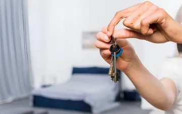 Hände halten einen Schlüsselbund einer Wohnung vor dem Hintergrund eines Schlafzimmers — Symbol der Wohnreform, deren sechswöchiges Frist abgelaufen ist, ohne dass der Kabinettsbeschluss untergeordnete Erlasse verabschiedet hat