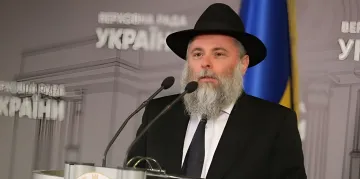 Главный раввин Киева в чёрном костюме и шляпе с длинной седой бородой выступает у трибуны с микрофонами на фоне украинского флага и надписи УКРАЇНА, заявив о готовности стать поручителем Ирины Мудрой в ВАКС