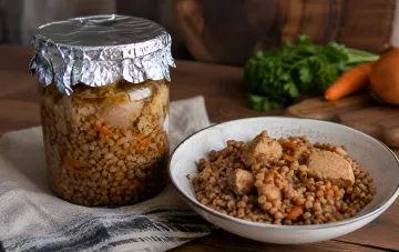 Домашняя заготовка на зиму: каша с мясом и морковью в стеклянной банке, закрытая фольгой, и порция гречки с кусочками мяса в тарелке