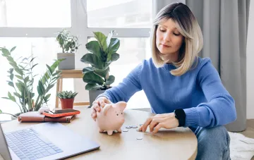 Frau legt Münzen in ein Sparschwein am Schreibtisch mit Laptop, illustriert wie man Ersparnisse vor Inflation schützt laut neuer Makroprognose der NBU