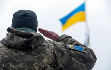 Украинский военный в камуфляже отдает салют украинскому флагу на фоне новости об упрощенном возврате из СЗЧ
