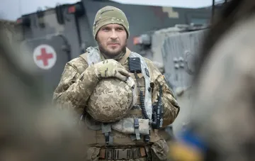 Ukrainian soldier in camouflage holding a helmet in front of military vehicles during counteroffensives in Sumy and Kharkiv regions