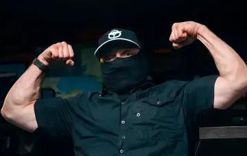 Commander of the Black Sea Legion of the GUR in a mask and cap with the unit emblem flexes in a pose of readiness against a map backdrop