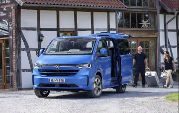 Синий Volkswagen Caravelle — семейный минивэн с открытой сдвижной дверью и багажником, пара с чемоданами у отеля