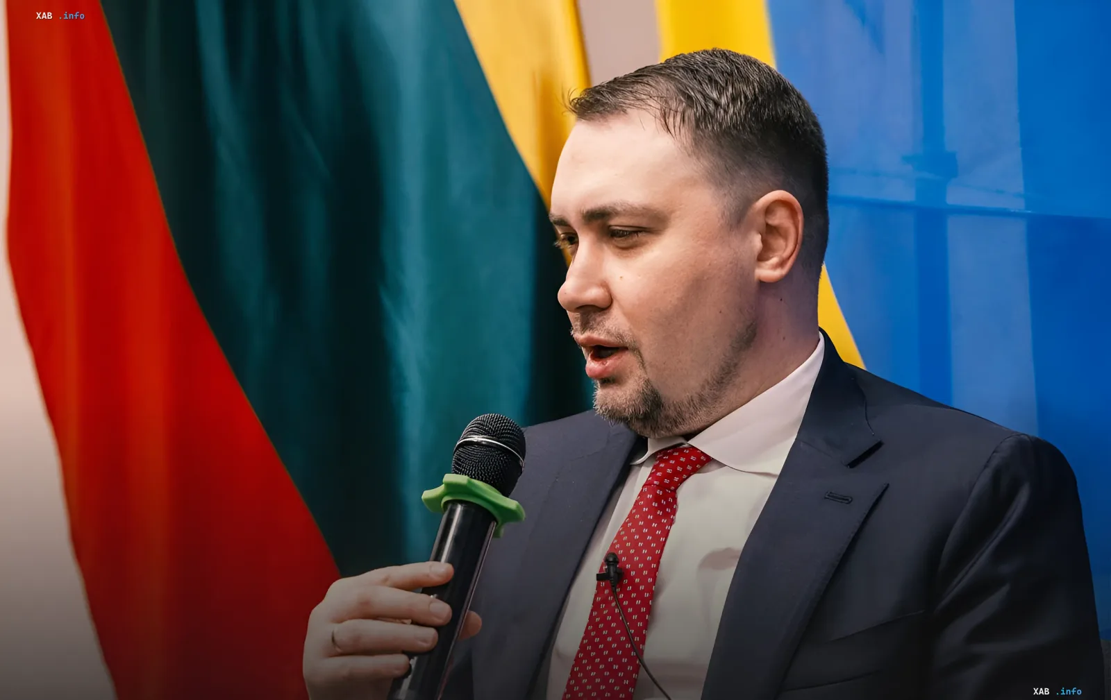Кирилл Буданов выступает с микрофоном на фоне государственных флагов, комментируя протесты в Украине 2025 года