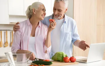 Reifes Paar in der Küche, das einen gesunden Salat aus frischem Gemüse zubereitet und ein Rezept auf dem Laptop ansieht – die Rolle der Essgewohnheiten für die Gesundheit