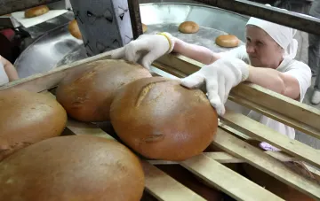 A baker in white headwear and gloves arranges freshly baked round loaves on bakery shelves — bread production continues without disruption
